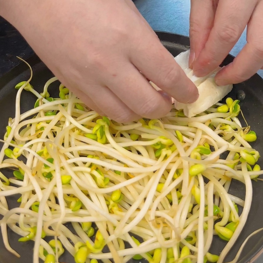 Manos colocando cuidadosamente rebanadas gruesas de tofu blanco sobre una cama de brotes de soja en una olla negra poco profunda.