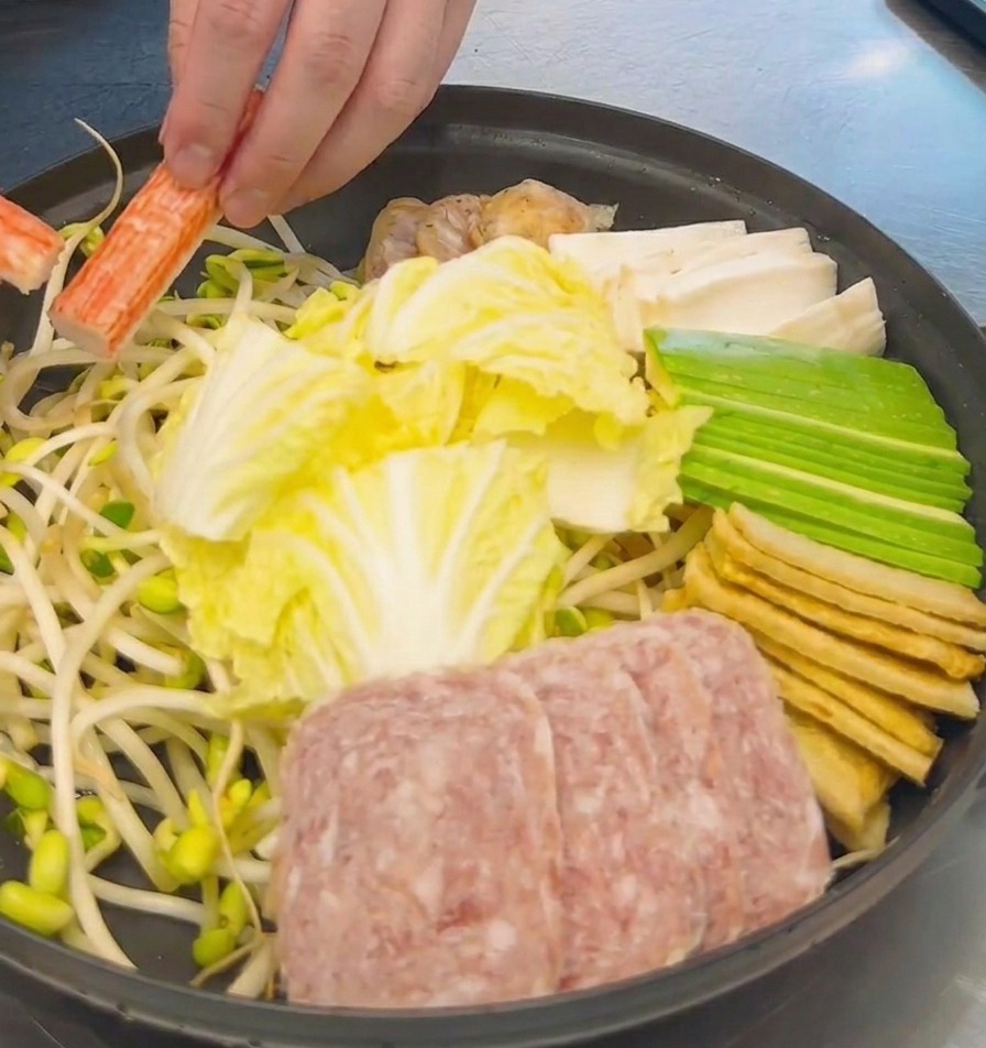 Una mano acomodando palitos de cangrejo naranjas y blancos en una olla ya llena de col cortada, spam y tofu.