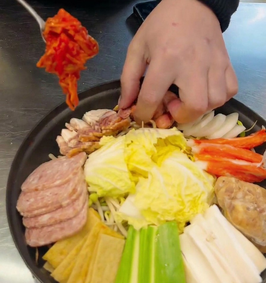 Una mano usando una cuchara de metal para colocar una porción de kimchi rojo en una olla poco profunda ya llena de salchichas, spam y verduras.