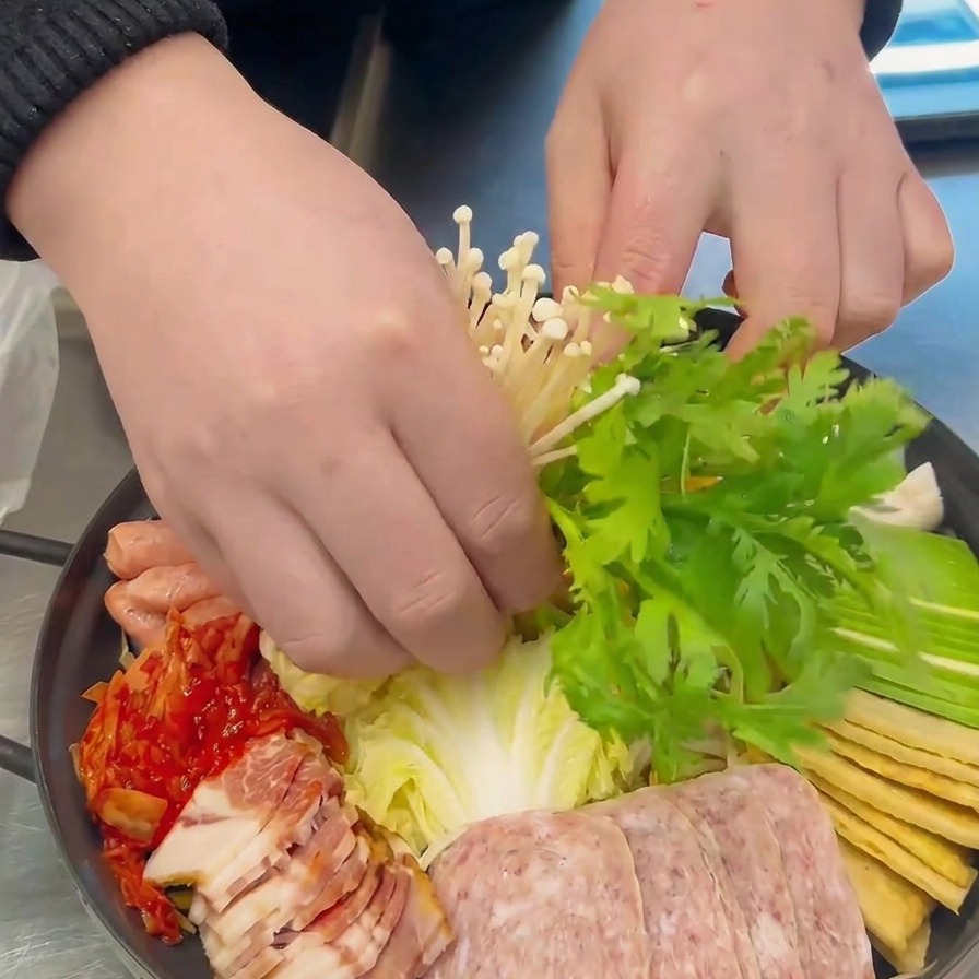 Manos colocando cuidadosamente un racimo de setas enoki blancas y hojas verdes de crisantemo en el centro de la olla preparada.