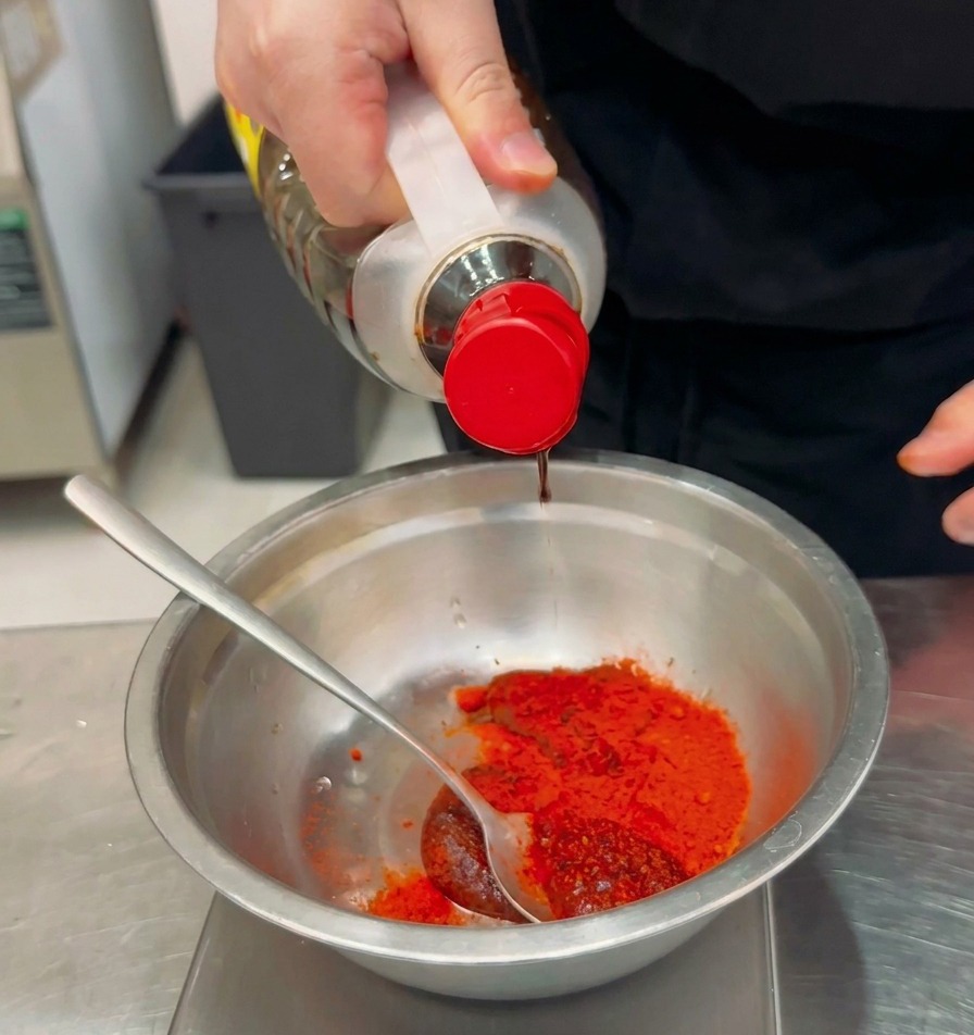 Condimento líquido siendo vertido desde una botella transparente en un bol de metal que contiene chile rojo en polvo y pasta de chile.