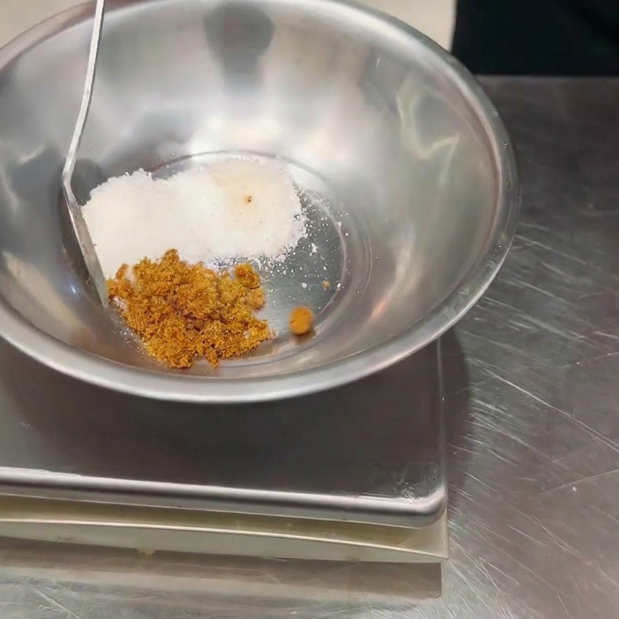 Una mano usando una cuchara para añadir condimento en polvo marrón a un tazón de metal que contiene polvo blanco, colocado sobre una báscula digital.