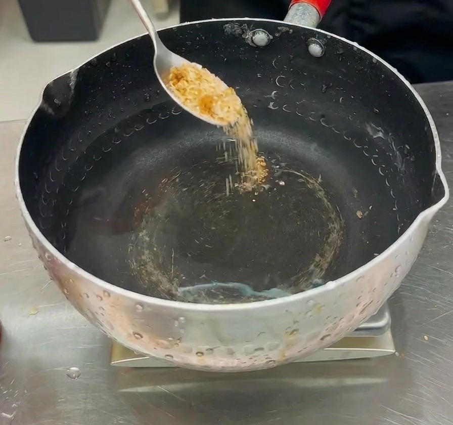 Polvo sazonador siendo vertido desde una cuchara en una olla de agua clara para preparar el caldo del estofado.
