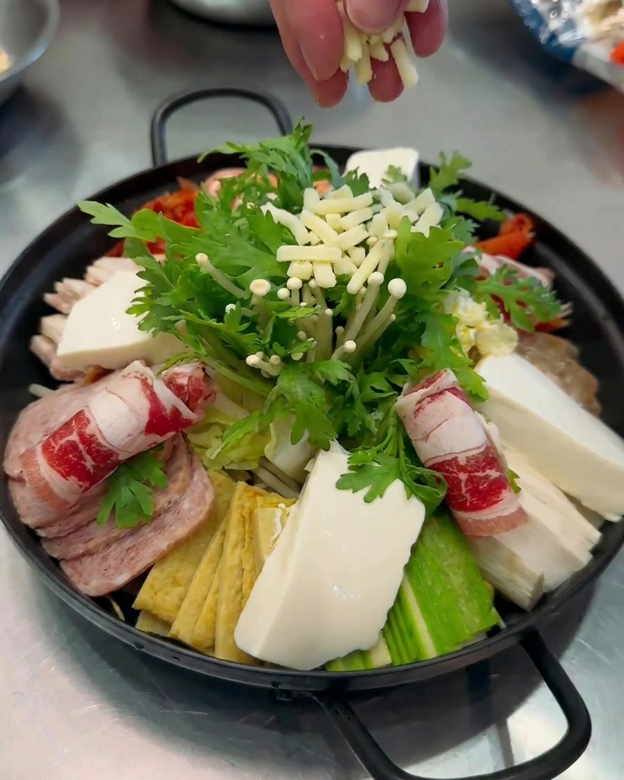 Primer plano de queso blanco rallado siendo espolvoreado a mano sobre una colorida olla de Budae-jjigae completamente armada.