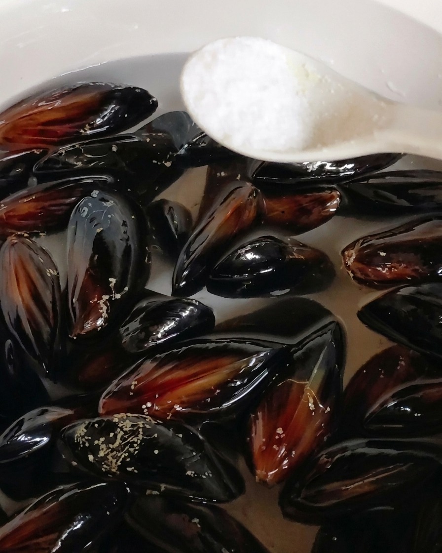 Una cucharada de sal siendo añadida a una olla con mejillones negros sumergidos en agua.