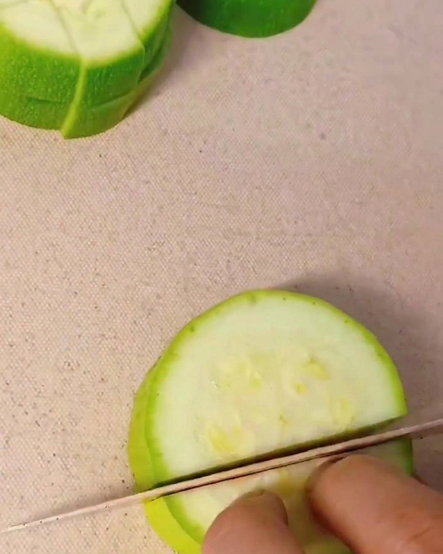 Primer plano de una mano usando un cuchillo de cocina para cortar un calabacín verde en dados pequeños sobre una tabla de cortar.