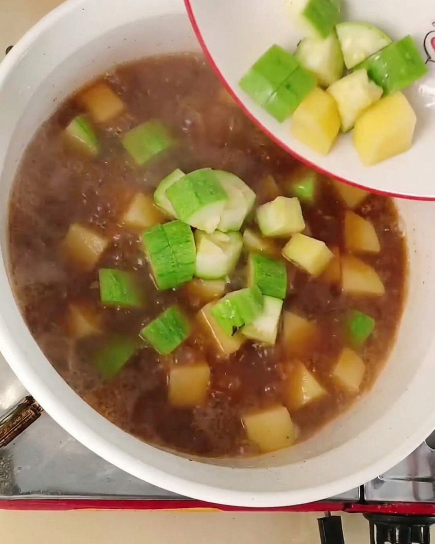 Dados de calabacín verde y cebolla blanca cayendo desde un plato blanco a un caldo rojizo hirviendo en una olla.