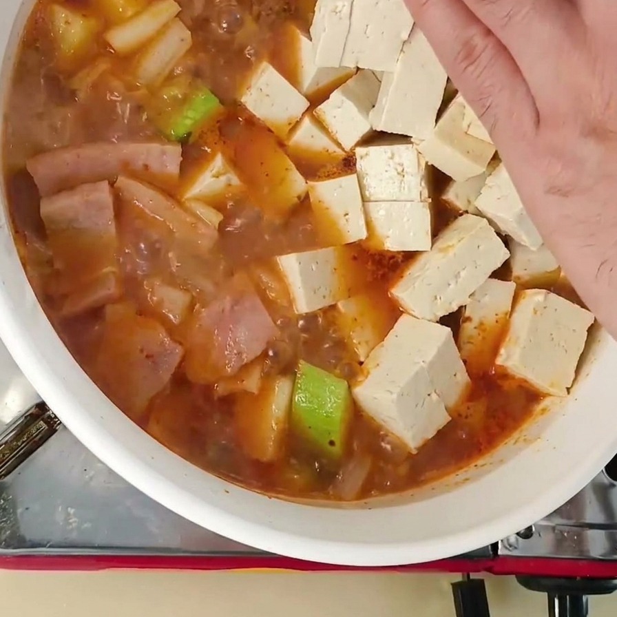 Una mano colocando cubos de tofu blanco fresco en una olla de estofado coreano hirviendo.