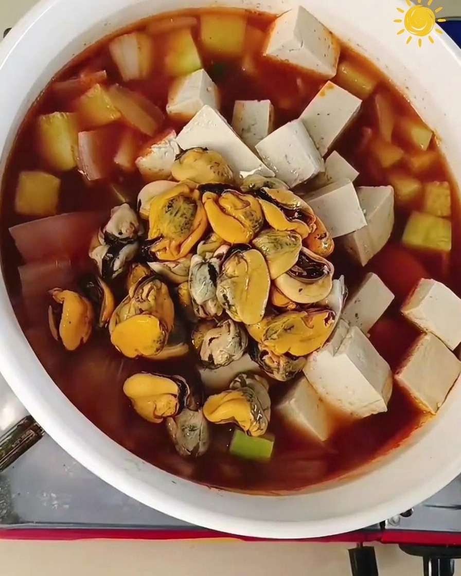 Carne de mejillón y varios langostinos crudos grandes siendo añadidos al centro de una olla hirviendo con tofu y verduras.