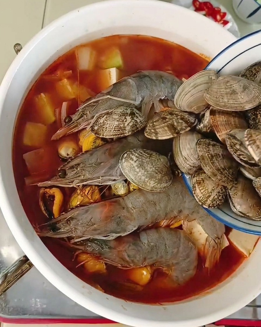 Un pequeño cuenco vertiendo almejas frescas enteras en una olla con langostinos, mejillones y tofu en un caldo rojizo.