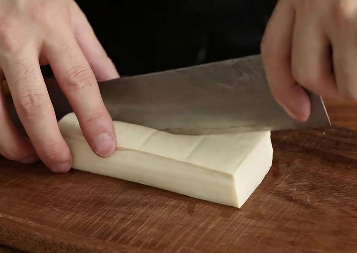 Una mano con un cuchillo de chef cortando un bloque de tofu blanco en cubos pequeños sobre una tabla de madera oscura.