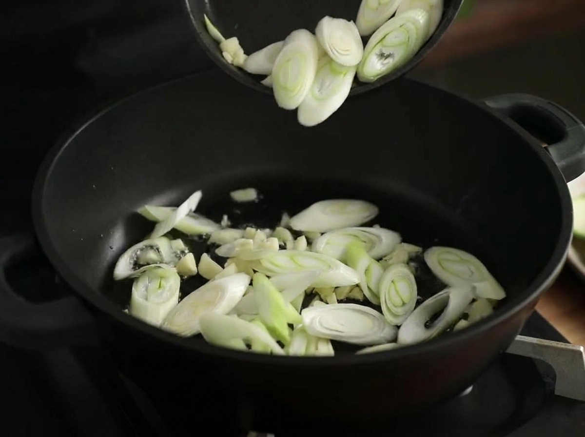 Ajo picado y rodajas de puerro salteándose en una olla negra con aceite caliente.