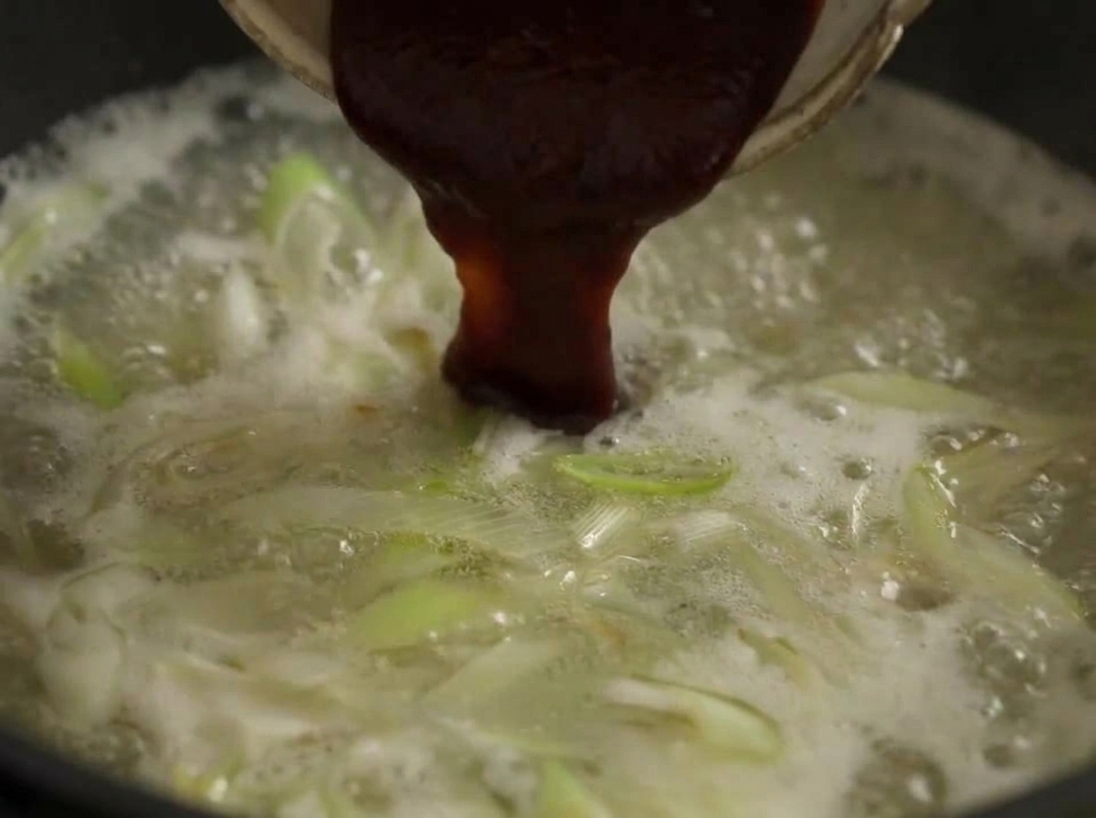 Una cuchara grande de pasta de chile rojo oscuro mezclándose en una olla de caldo hirviendo con puerros.