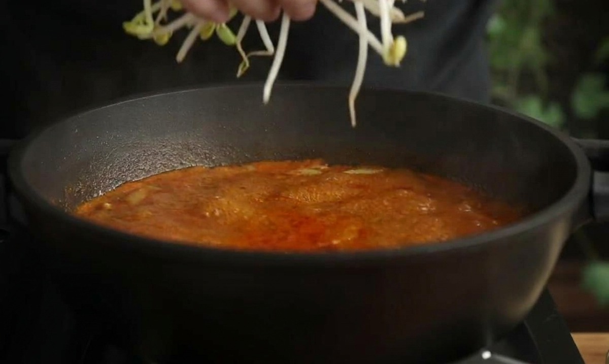 Una mano añadiendo un puñado de brotes de soja largos en una olla de sopa de mariscos roja hirviendo.