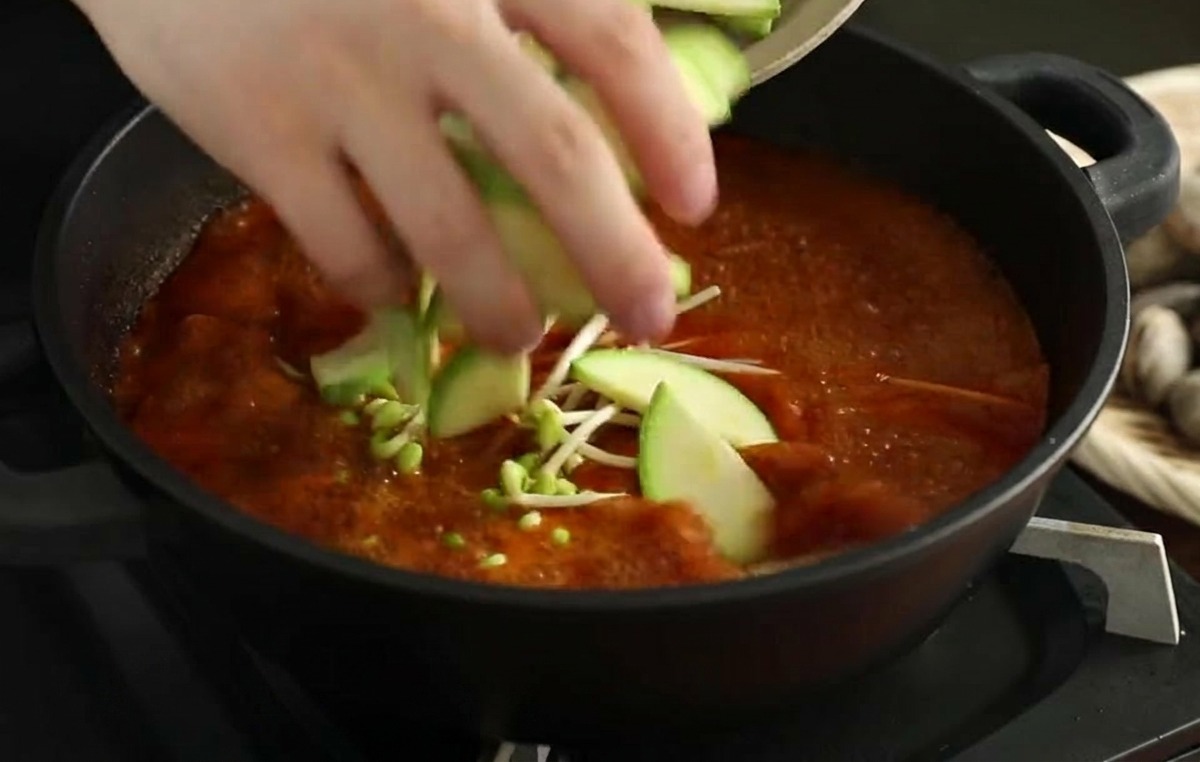 Manos colocando rodajas de calabacín fresco en un caldo de mariscos rojo que ya contiene brotes de soja.