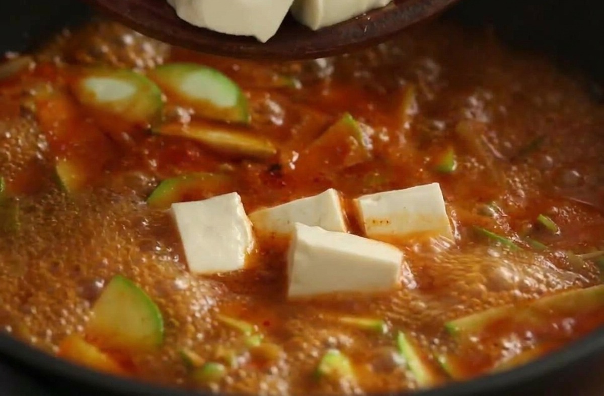 Cubos de tofu blanco cayendo desde un tazón a un vibrante estofado coreano rojo.
