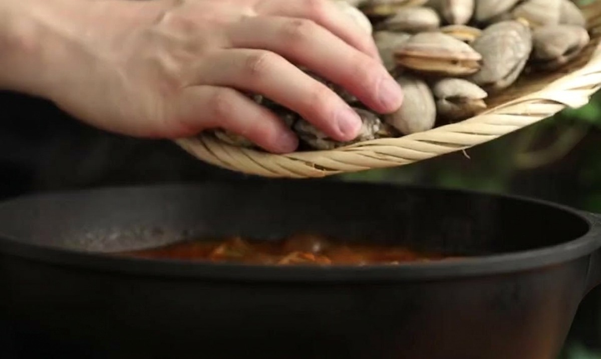 Una persona usando un colador de bambú para bajar almejas frescas en una olla de estofado rojo.