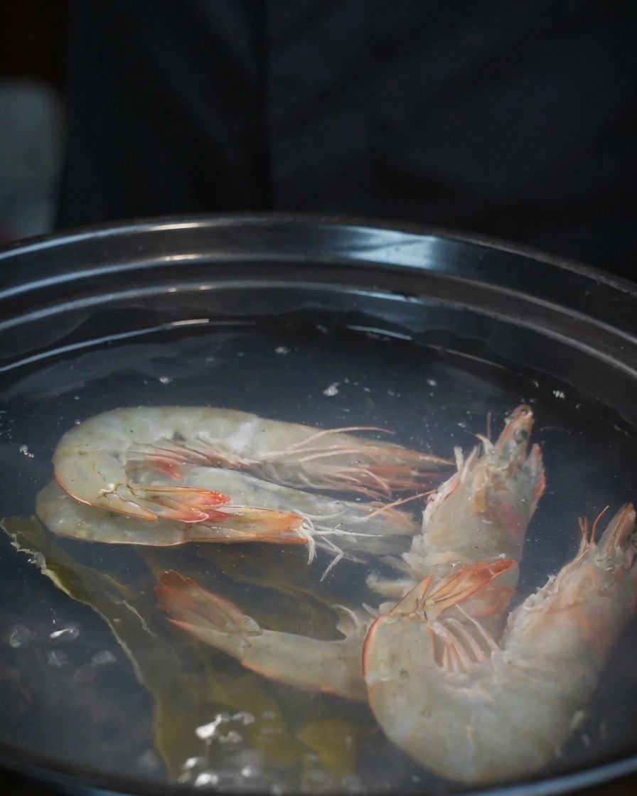 Almejas frescas cayendo desde un cuenco a una olla que ya contiene camarones y caldo.