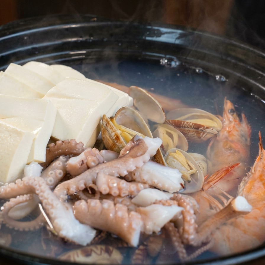 Una olla de barro con almejas y camarones donde se añaden rodajas de rábano blanco y zanahoria naranja con una espátula azul.