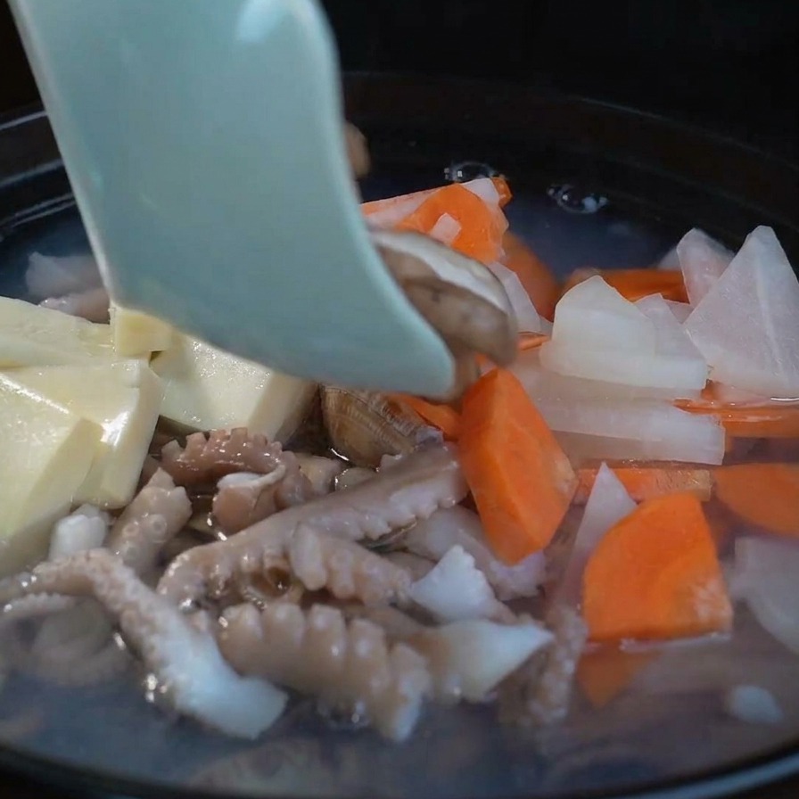 Rodajas de setas shiitake frescas siendo añadidas a una sopa de mariscos burbujeante junto al tofu y los vegetales.