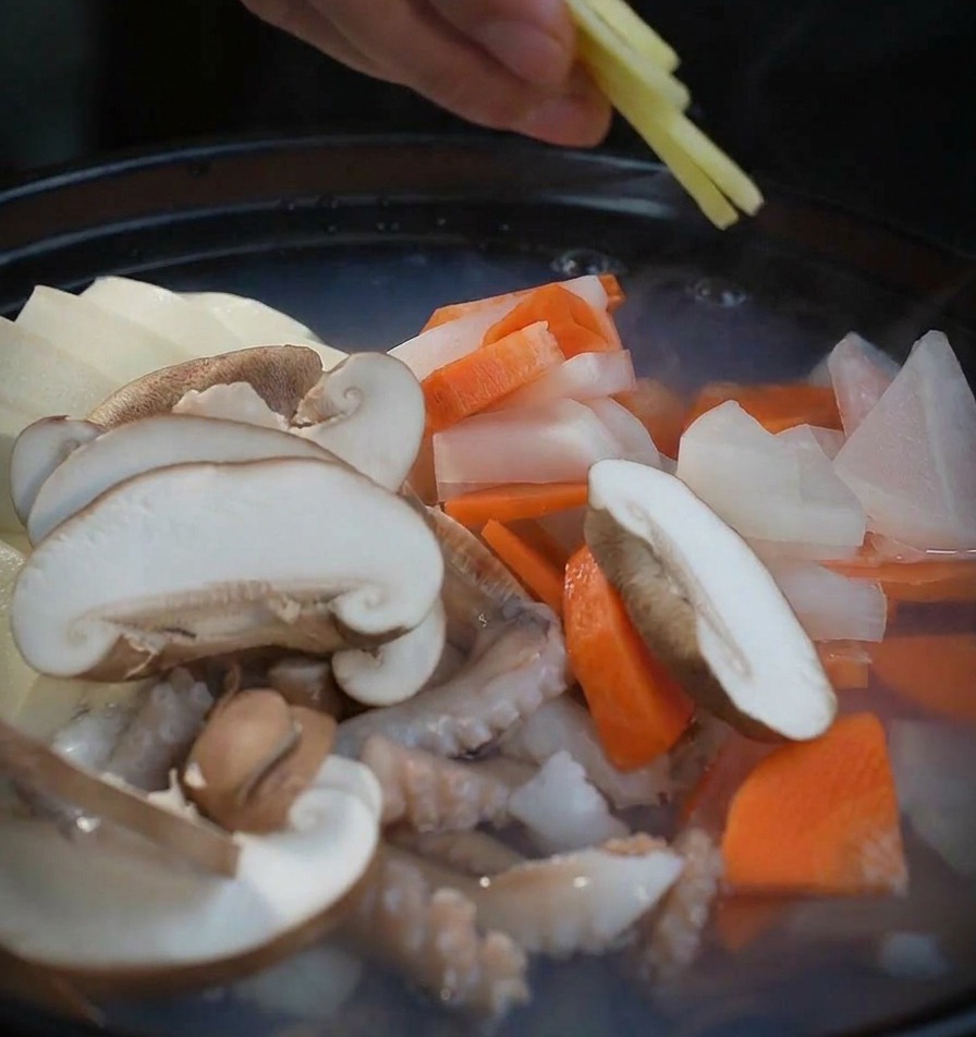 Una mano colocando rodajas finas de jengibre fresco en una olla hirviendo llena de setas, rábano y mariscos.