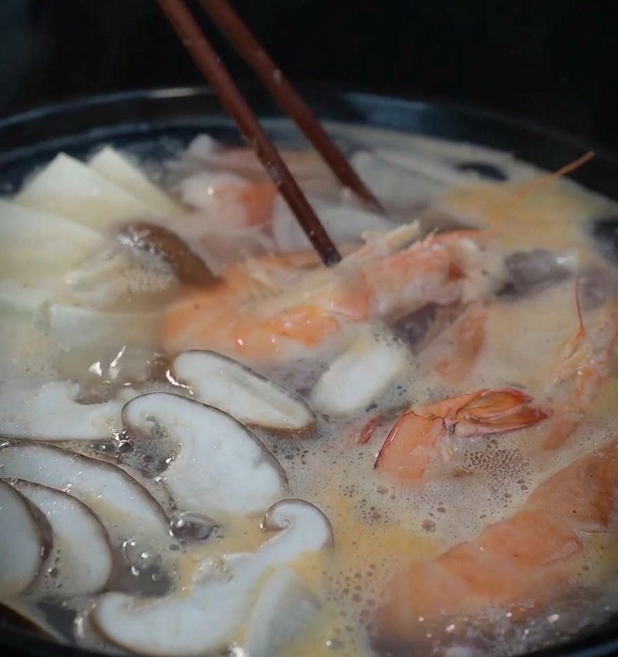 Una olla negra humeante con camarones, setas y tofu hirviendo en un caldo ligero, removido con palillos de madera oscuros.
