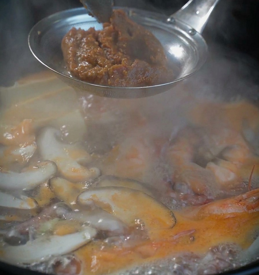 Una cucharada de pasta de soja marrón y espesa en un colador de metal sobre una olla de sopa de mariscos burbujeante.