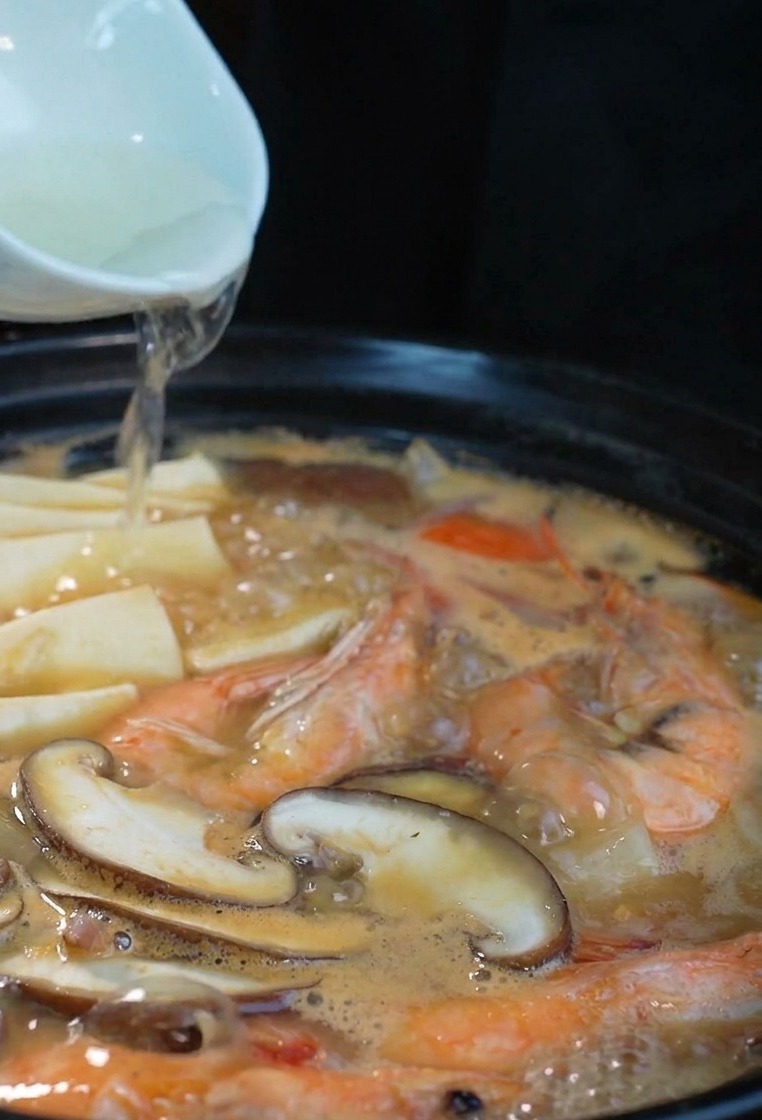 Un chorro de sake transparente vertido desde un cuenco de cerámica blanca en una olla negra humeante con mariscos.