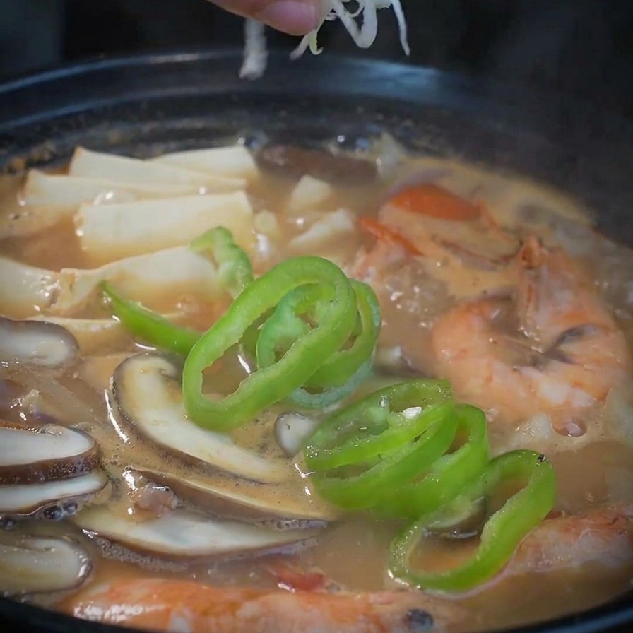 Una mano esparciendo una mezcla de cebolleta blanca y verde picada sobre una olla humeante de sopa terminada.