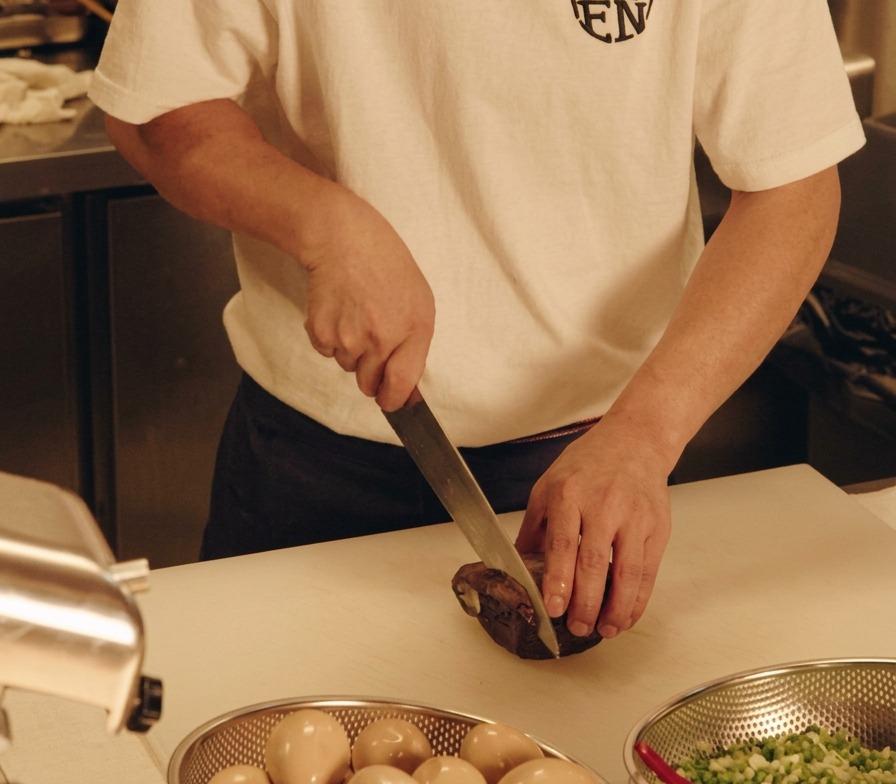 Un chef cortando una pieza de panceta de cerdo estofada en rodajas gruesas sobre una tabla de cortar blanca.