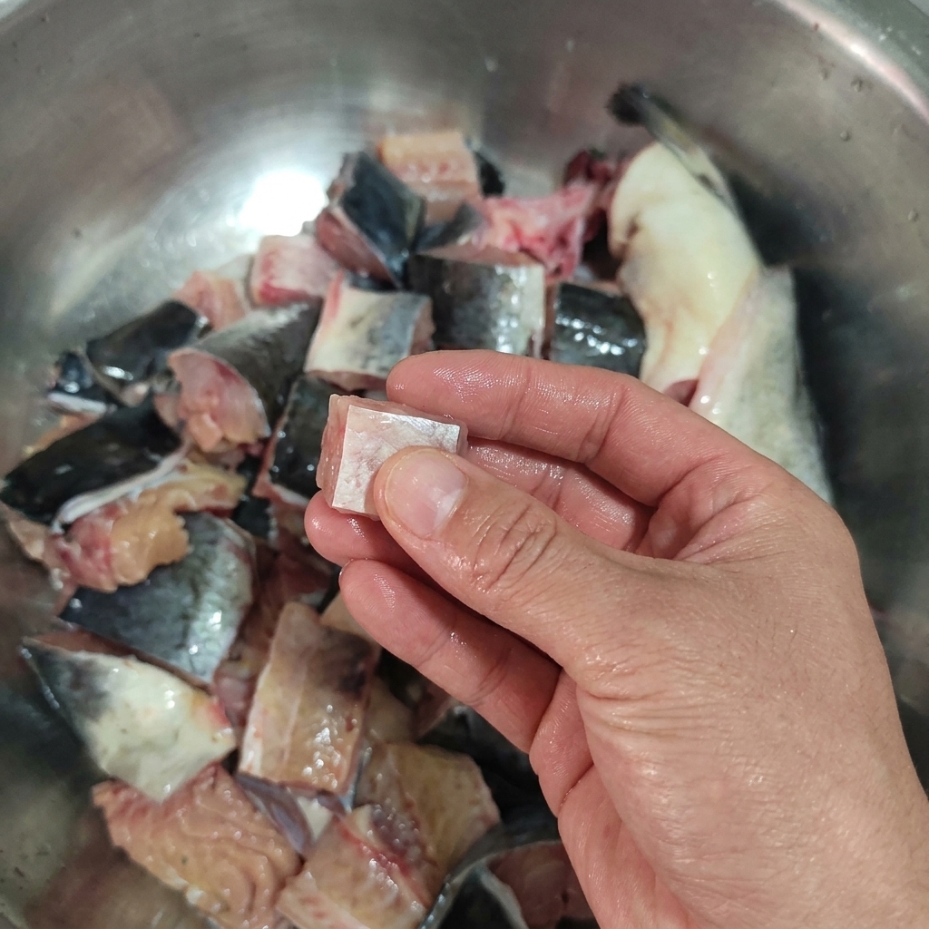 Una mano sosteniendo un trozo cuadrado de bagre crudo frente a un bol con más trozos cortados.