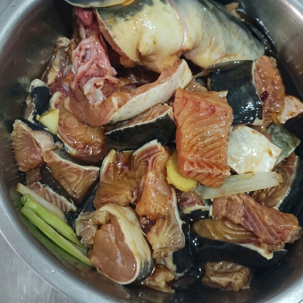 Trozos de pescado crudo mezclados con jengibre, cebolleta y un marinado oscuro en un bol de metal.