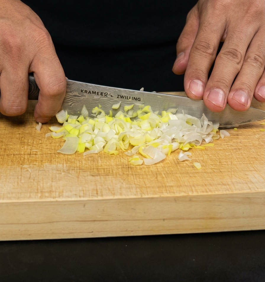 Manos de una persona usando un cuchillo de chef para picar finamente cebolletas sobre una tabla de madera clara.