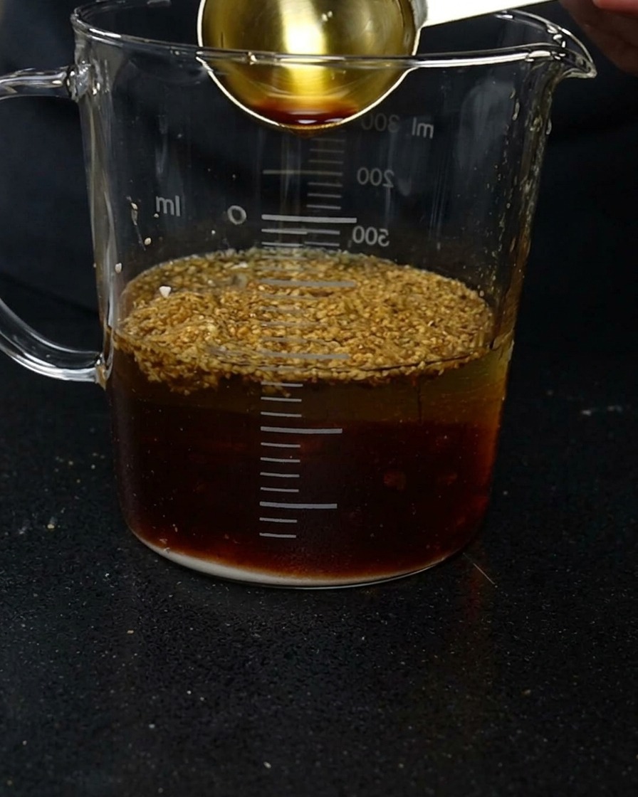 Una cuchara medidora dorada vertiendo salsa de soja oscura en una jarra de cristal con agua y varios condimentos en polvo.