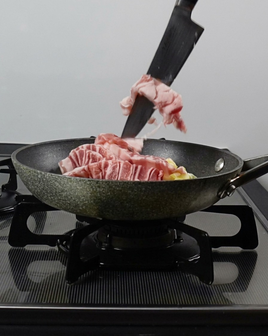 Un cuchillo empujando tiras de panceta cruda hacia una sartén que ya tiene cebolleta picada y ajo.