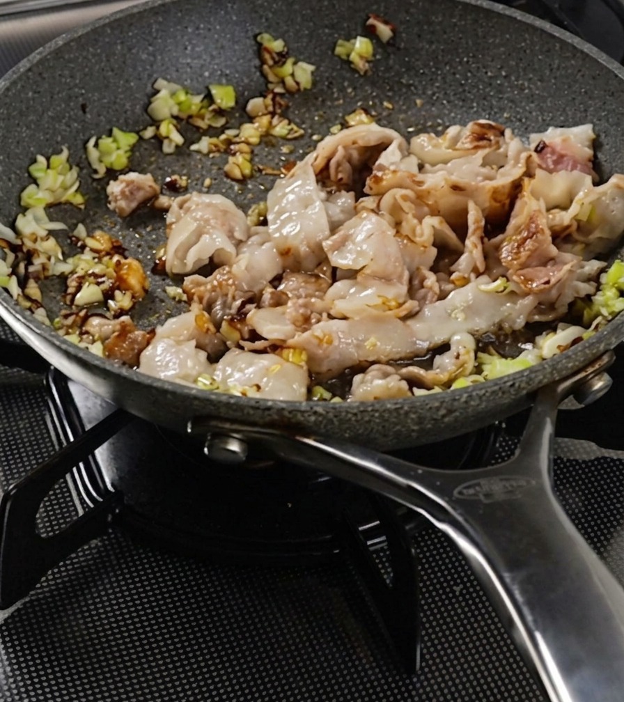 Tiras de panceta y cebolleta salteándose en una sartén hasta que la superficie de la carne se ve dorada.