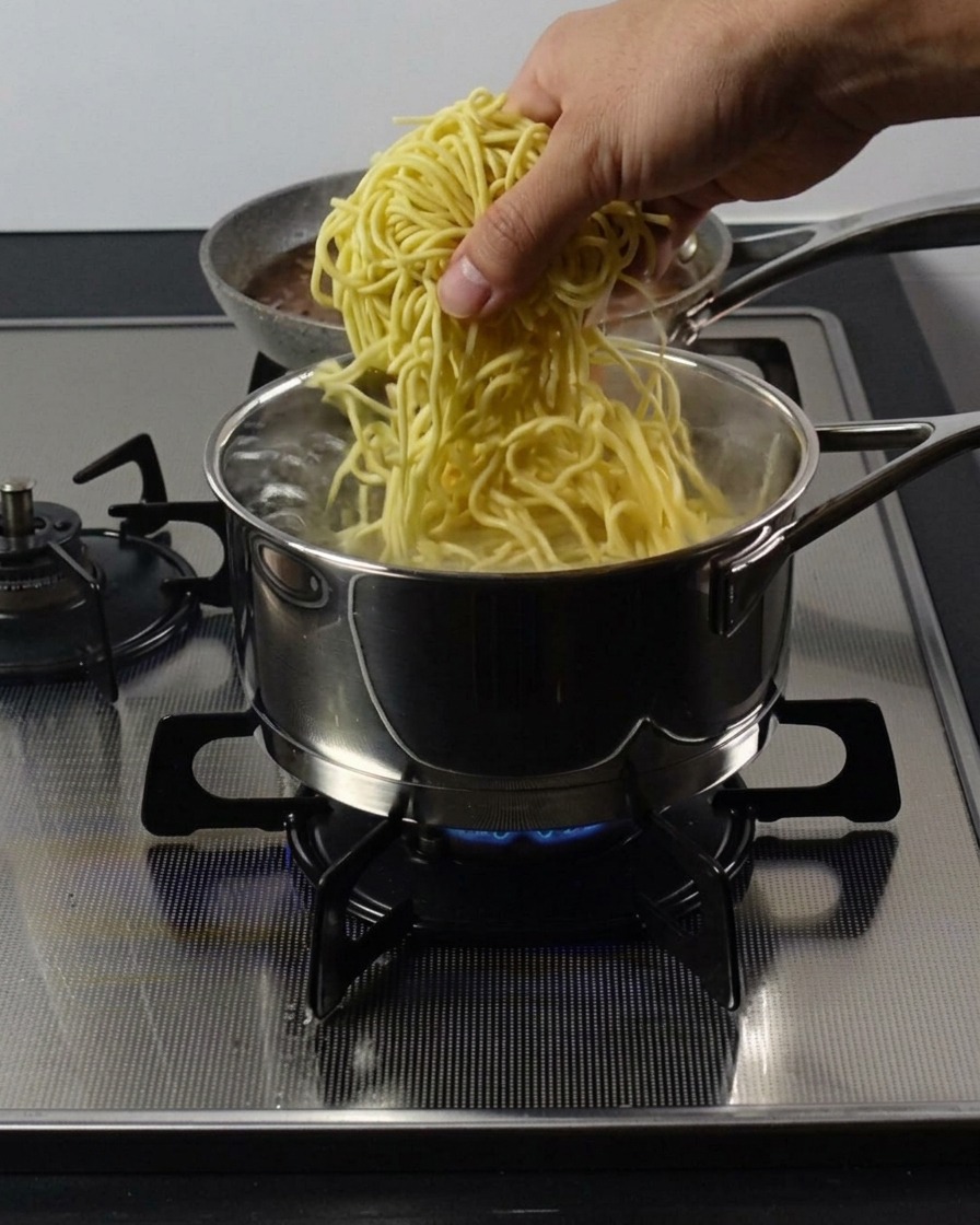 Una mano sumergiendo un nido de fideos ramen gruesos en una olla de acero inoxidable con agua hirviendo.