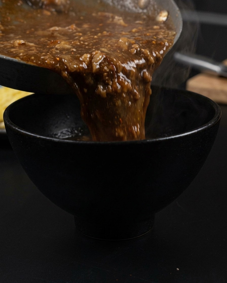 Caldo oscuro y espeso con trozos de cerdo vertiéndose de una sartén a un bol negro mate, con vapor saliendo del líquido.