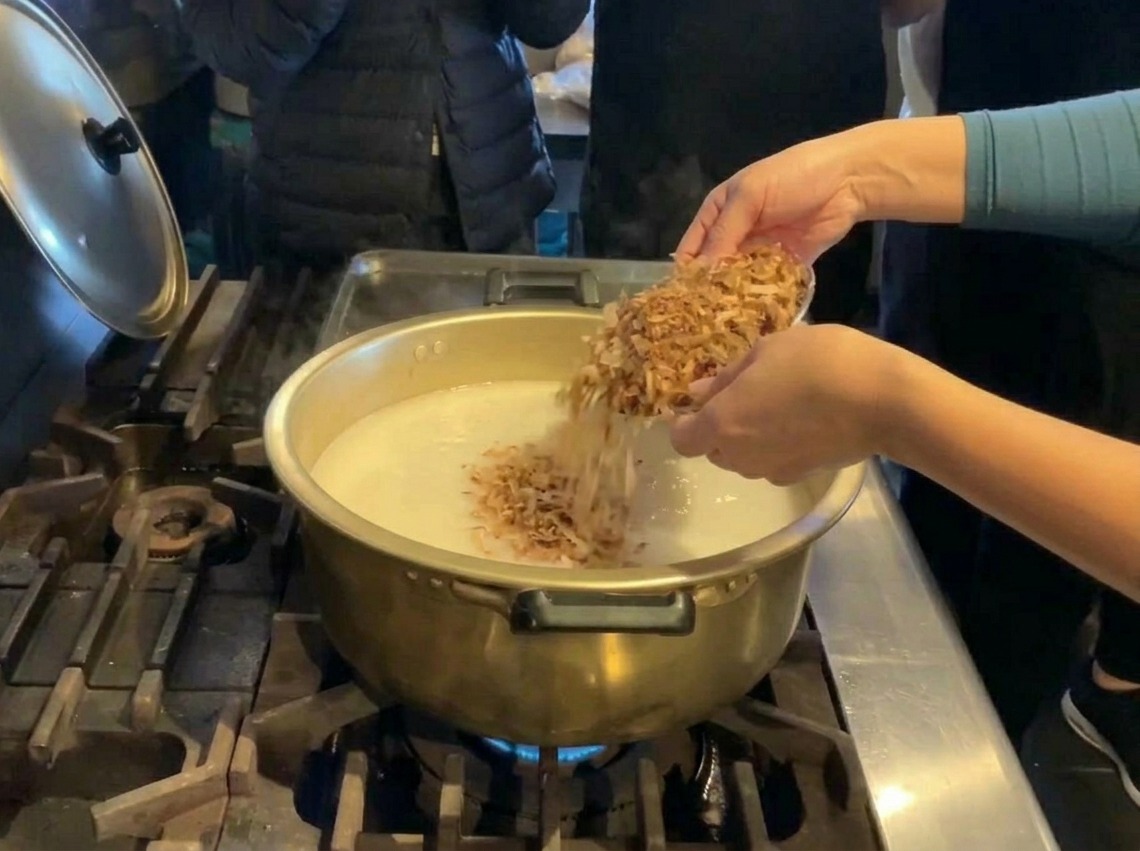 Una persona vertiendo una gran cantidad de copos de pescado seco desde un bol plateado en una olla dorada con caldo blanco.