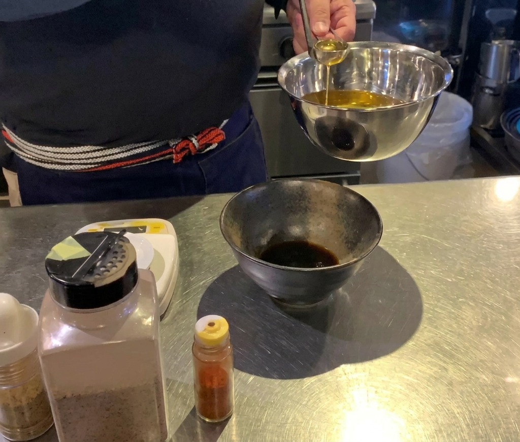 Un cocinero usando un pequeño cucharón de plata para verter aceite dorado en un cuenco negro con salsa oscura.