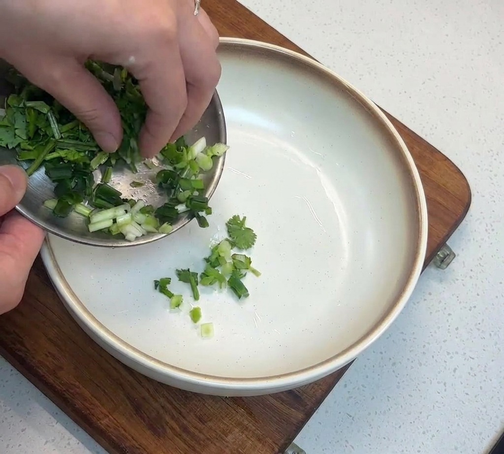 Primer plano de cebolletas verdes picadas siendo añadidas a un cuenco blanco sobre una tabla de madera.