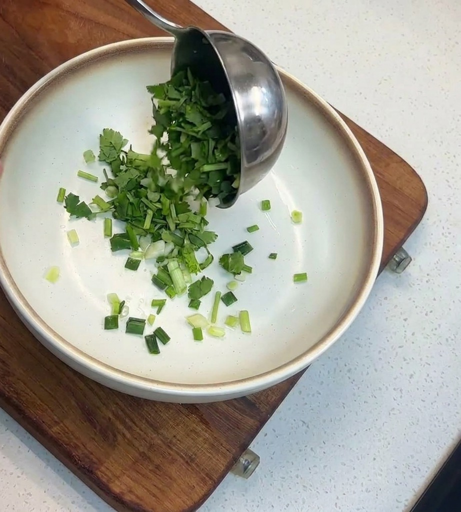 Cucharón vertiendo cilantro y cebolletas picadas en un cuenco blanco con borde marrón.