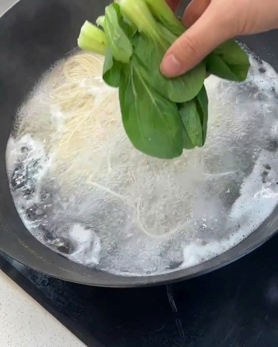 Mano echando hojas de bok choy fresco al wok con los fideos hirviendo.
