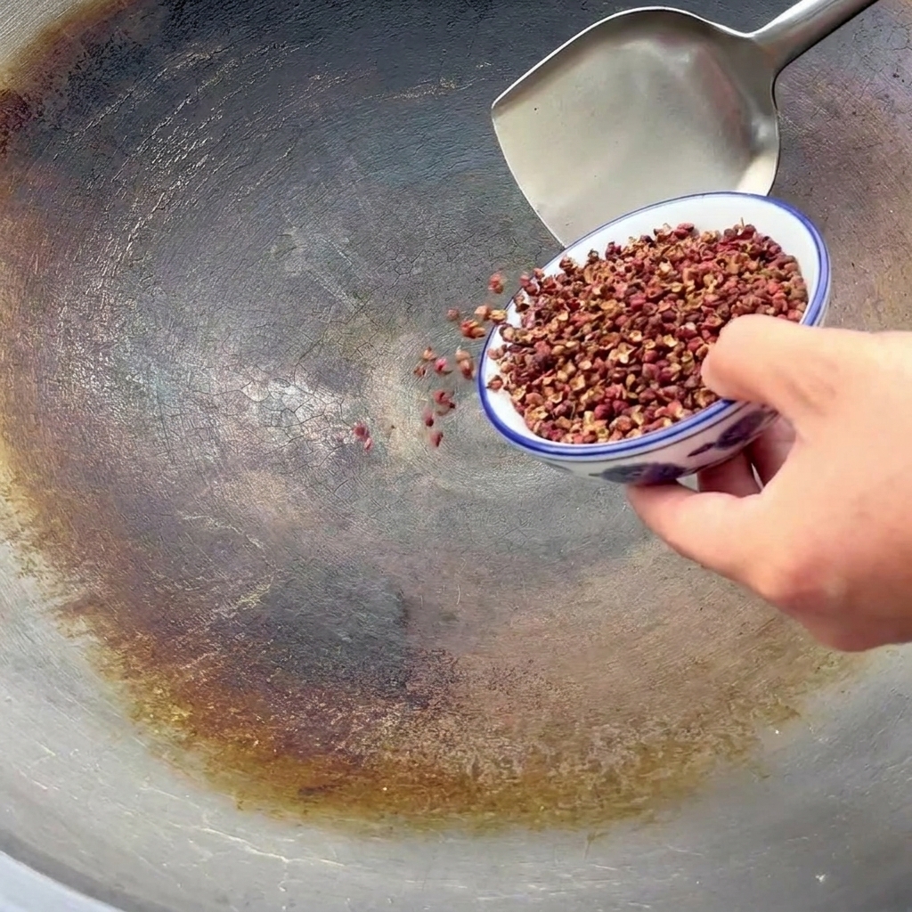 Pimienta de Sichuan vertiéndose desde un pequeño cuenco azul y blanco en un wok grande para tostar.