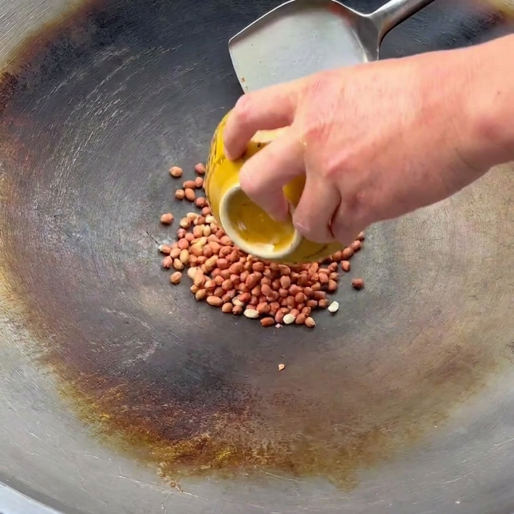 Una mano vertiendo cacahuetes crudos desde un cuenco amarillo en un wok donde una espátula de metal está lista para saltearlos.