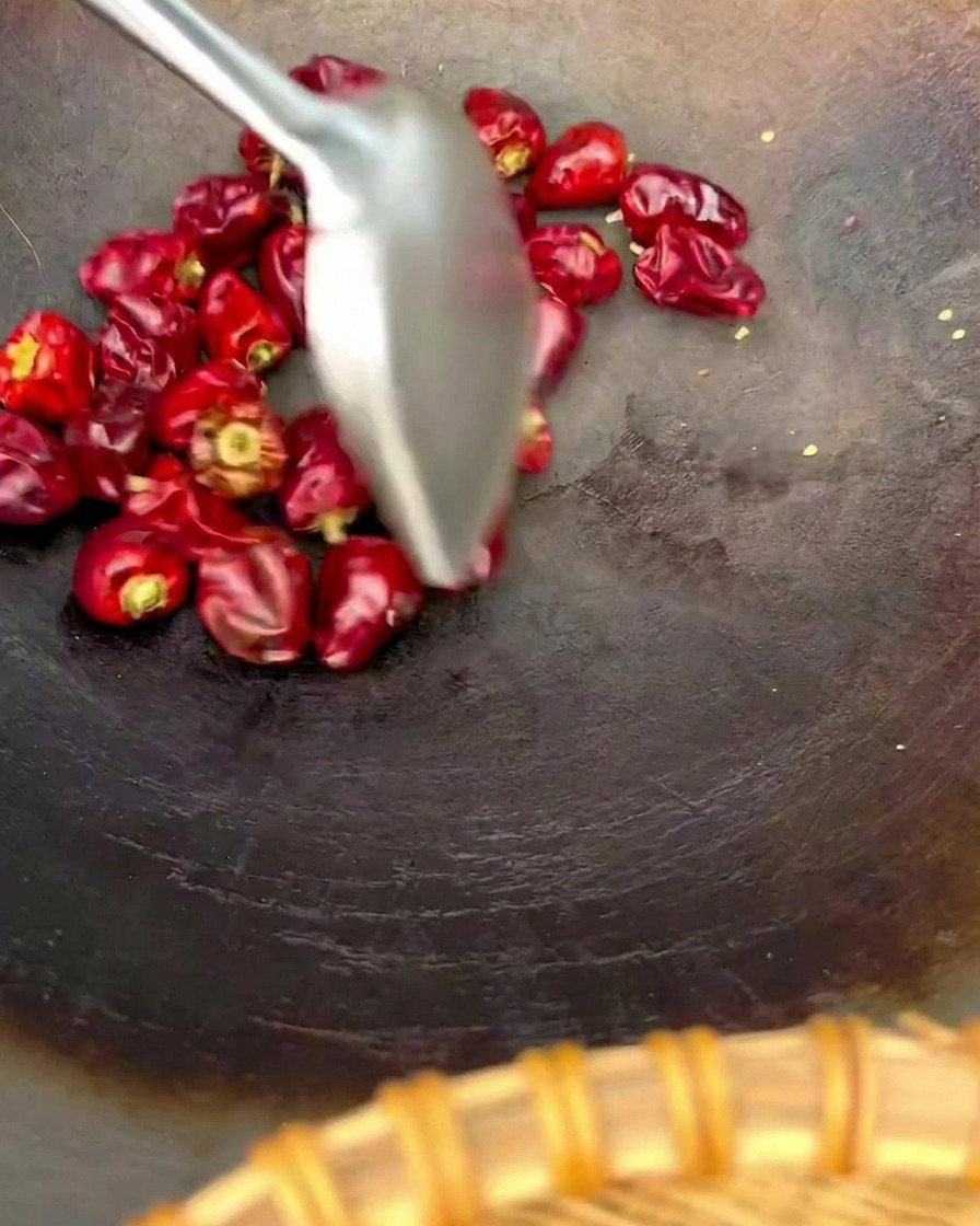 Una espátula de metal removiendo un puñado de chiles rojos redondos tipo linterna en un wok oscuro.