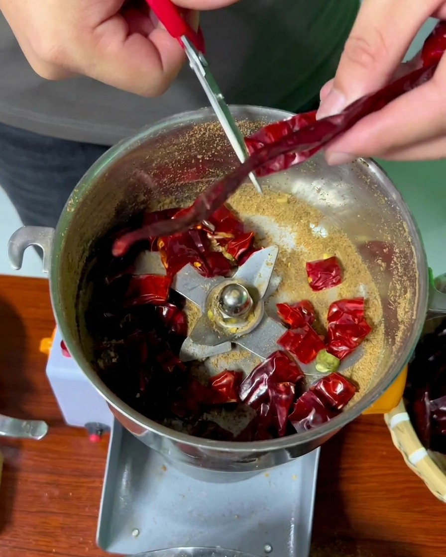 Manos usando tijeras de mango rojo para cortar chiles rojos secos en el recipiente de un molinillo de acero inoxidable.