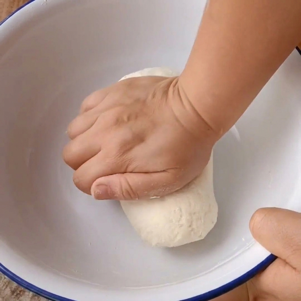 Dos manos amasando firmemente una bola de masa dentro de un bol blanco.