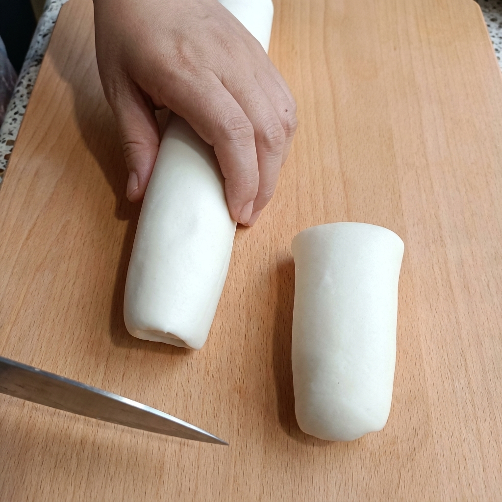 Una mano sosteniendo un cilindro de masa blanca en una tabla de cortar de madera mientras lo corta con un cuchillo.