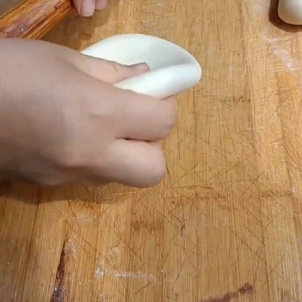 Una mano usando un rodillo de madera para aplanar un trozo de masa en un disco grueso sobre una tabla de madera.